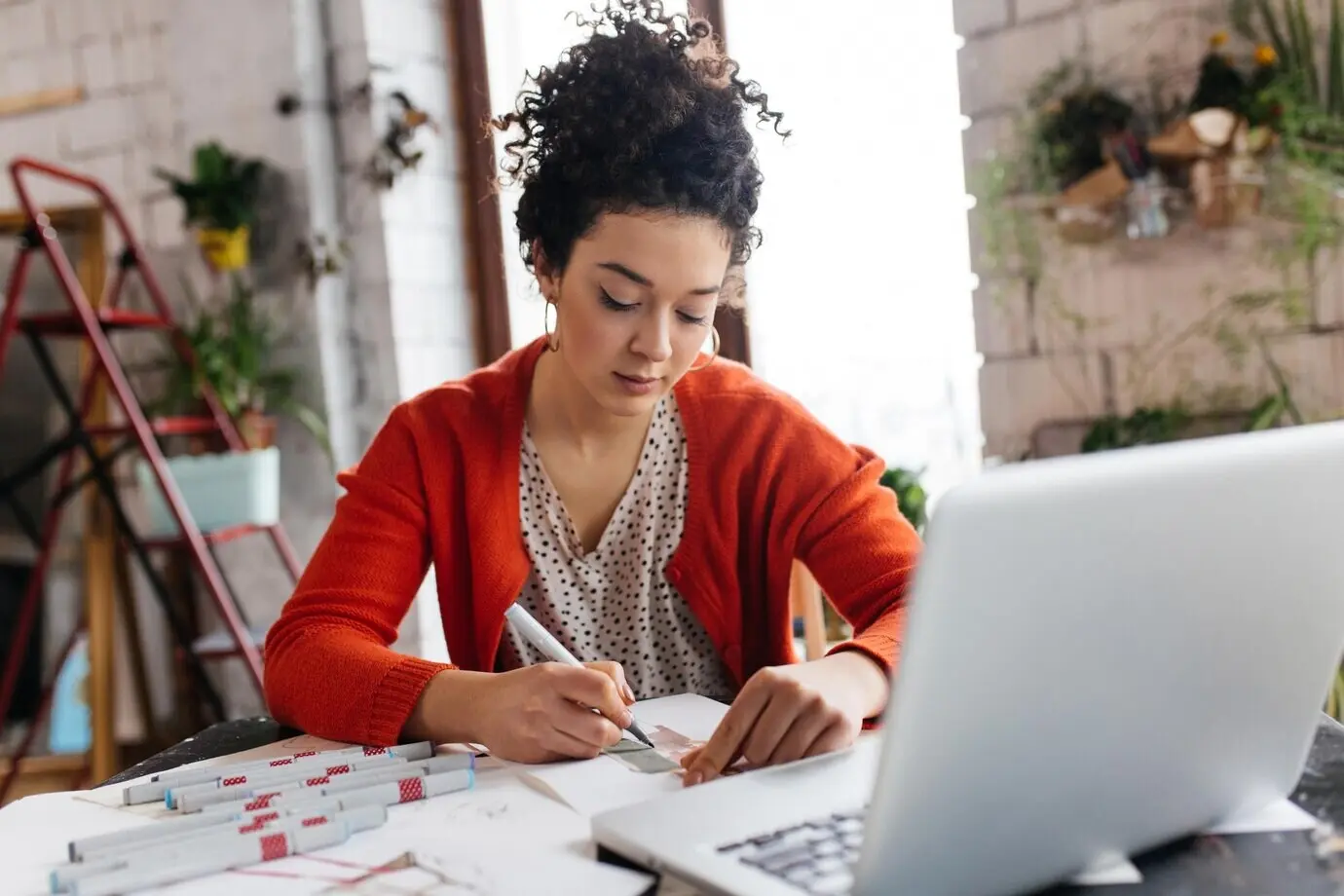 A young, beautiful woman with dark curly hair sits at a table with a laptop, dreamily sketching fashion illustrations while spending time in a modern, cozy workshop with big windows.
