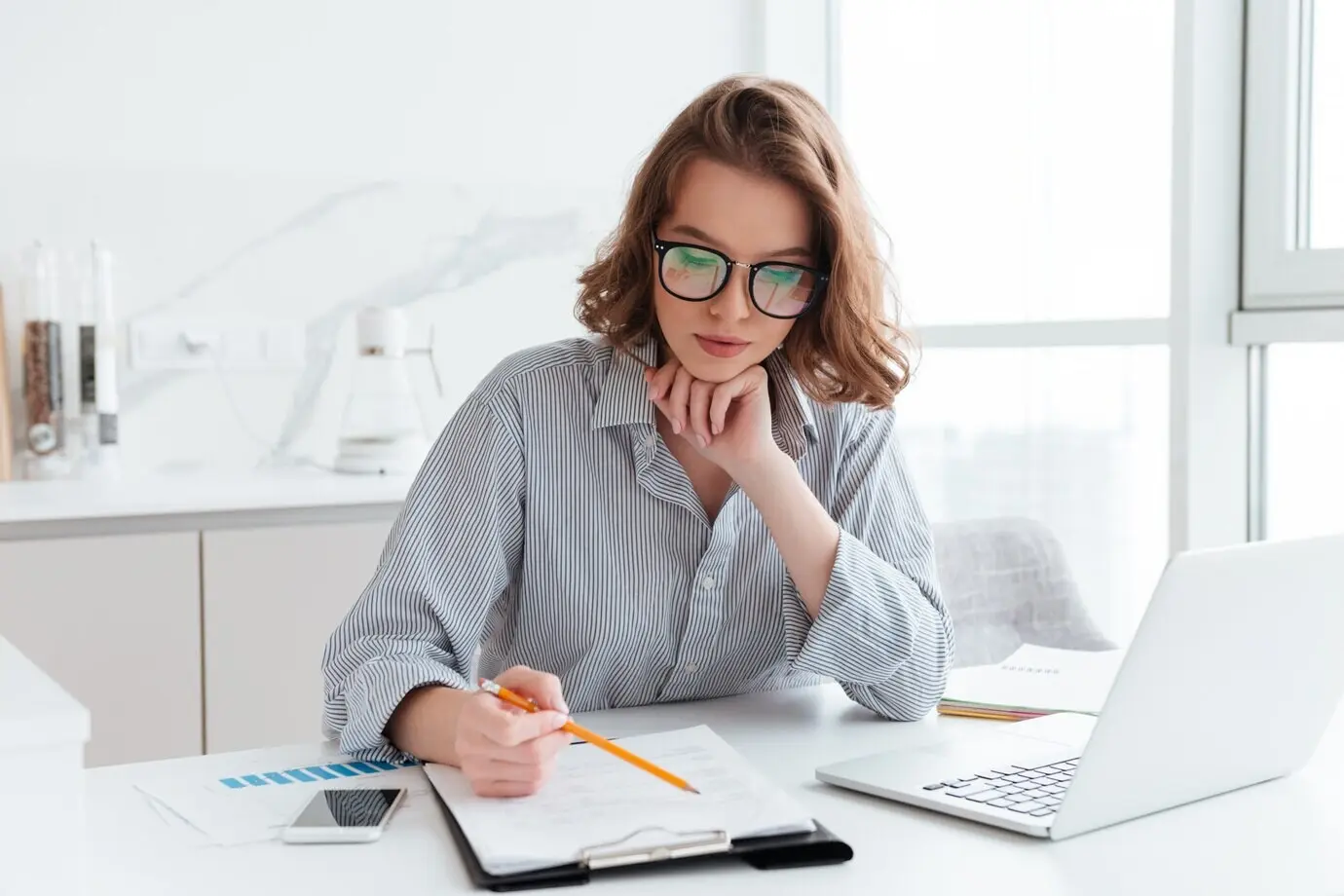 A young, focused businesswoman wearing glasses and a striped shirt works with papers at home.